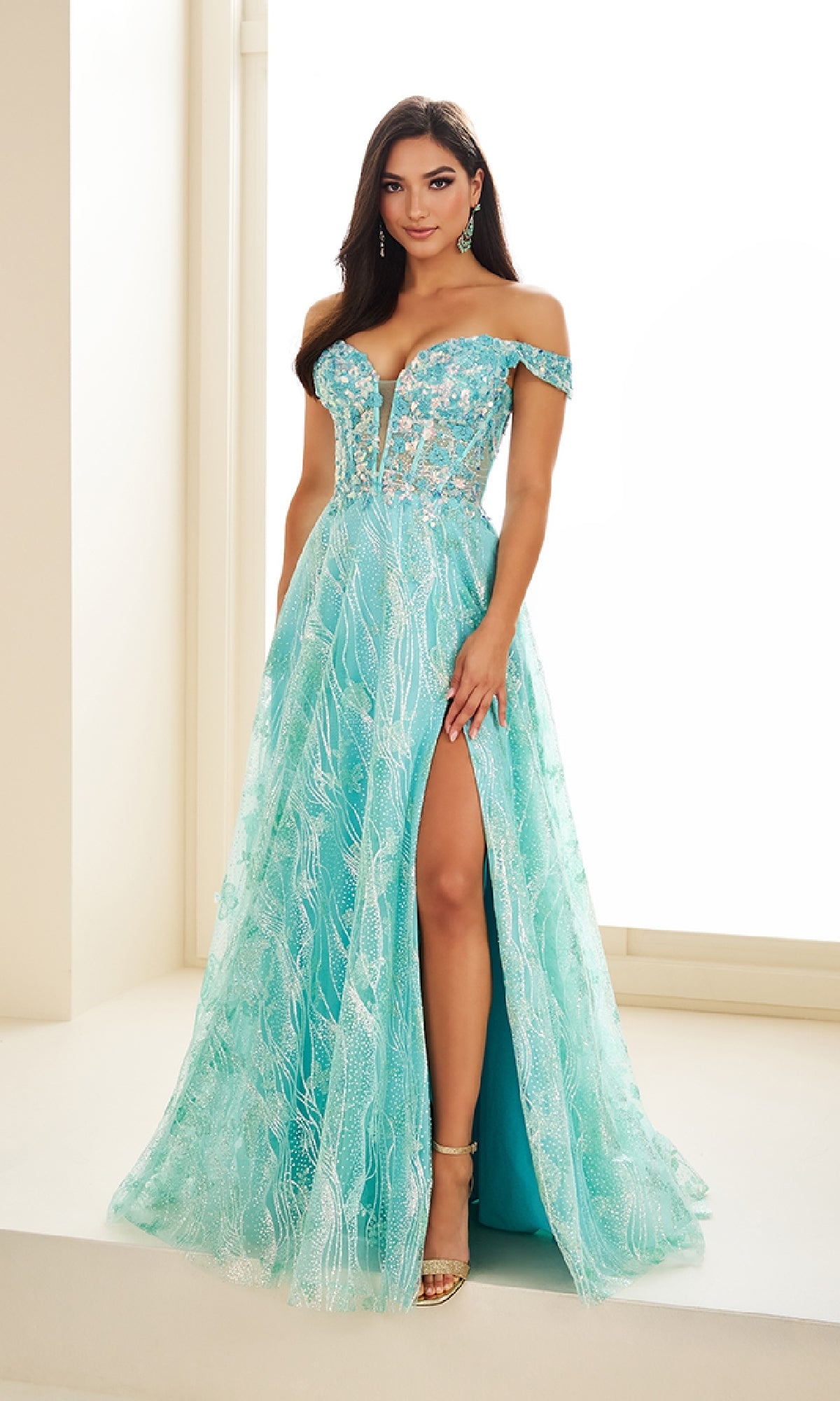 A woman stands indoors wearing the Glow-in-the-Dark Prom Dress: Ellie Wilde EW36039—an off-the-shoulder, floor-length aqua gown with floral embroidery, thigh-high slit, sheer details, and a figure-flattering corset bodice.